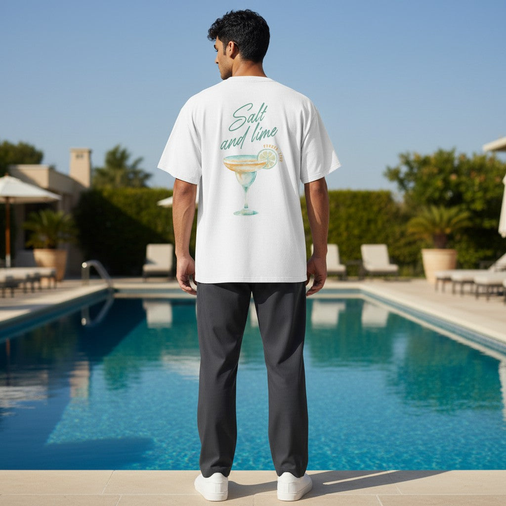 رجل يرتدي تيشرت أبيض بقصة واسعة من FLEXSTYLE يتميز بطبعة كوكتيل Salt and lime على الظهر بجانب مسبح خارجي.