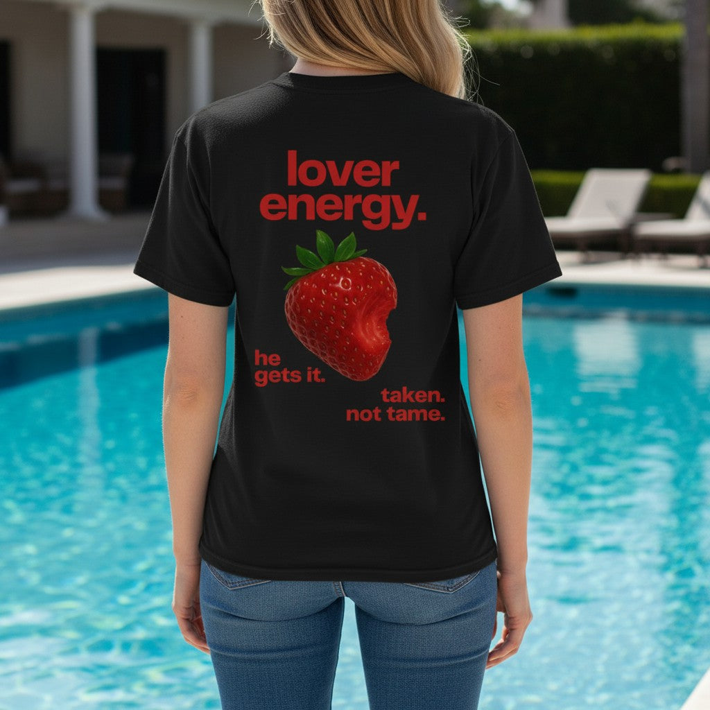 تيشيرت أسود بقصة واسعة من FLEXSTYLE يتميز بطبعة فراولة حمراء وعبارة lover energy على الظهر، ترتديه عارضة بجانب مسبح.